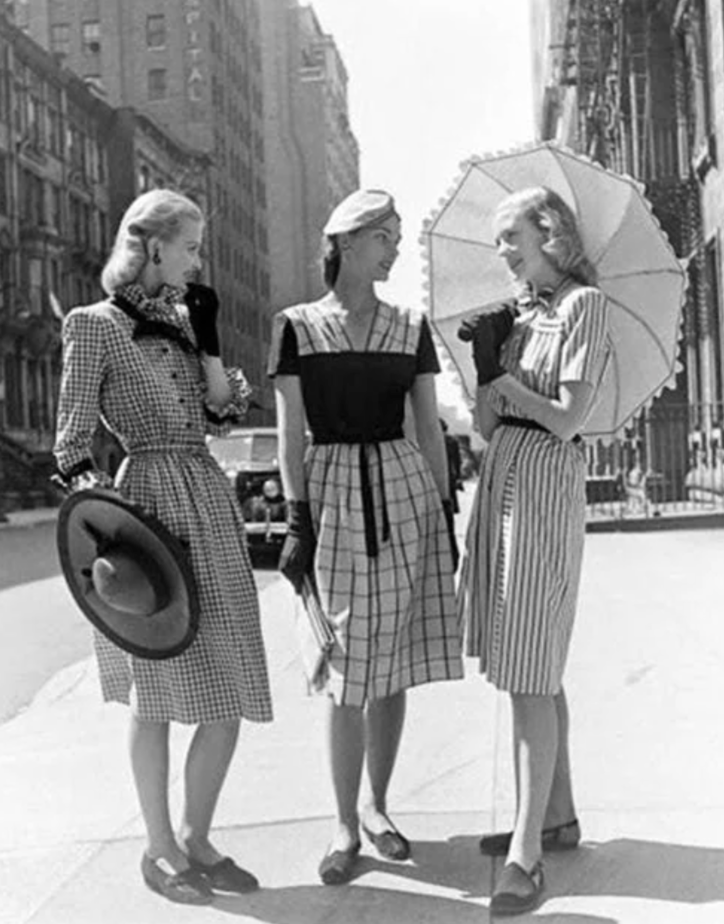 Three women in 1940s fashion stand on a city street. They wear dresses with belts and patterns, hats, and dark gloves. One holds a parasol. Behind them are tall buildings and a vintage car.