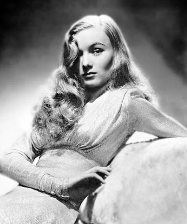 Black and white portrait of a woman with wavy hair wearing a shimmering long-sleeved dress. She is seated with one arm resting on a chair, looking toward the camera with a soft-focus background enhancing the vintage feel of the image.