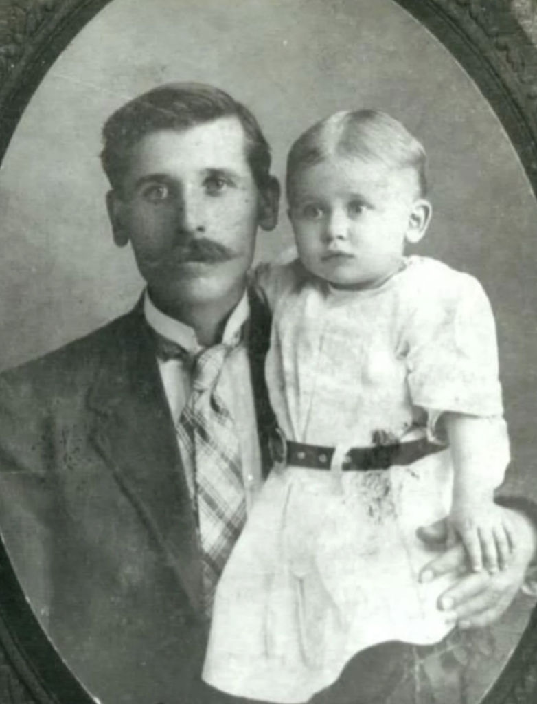 A black and white portrait of a man in a suit and tie holding a small child dressed in a light-colored outfit. The child's hair is neatly combed back. The image has an oval frame, giving it a vintage appearance.