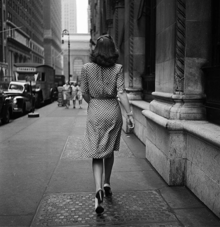 Woman in a polka dot dress walks down a city sidewalk lined with buildings. Vintage cars are parked along the street, and a group of people walks ahead. The setting appears to be a mid-20th century urban area.