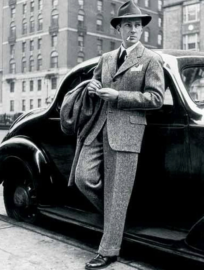 A man in a 1940s-style suit and hat leans against a vintage car, holding a coat. He stands on a city street with tall buildings in the background. The image is black and white, giving it a classic, timeless feel.