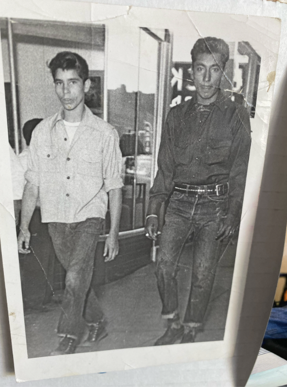 Black and white photo of two young men walking outdoors. One wears a light button-up shirt and jeans, the other a dark long-sleeve shirt and jeans. They appear to be on a sidewalk near storefronts. The photo looks vintage with some creases.