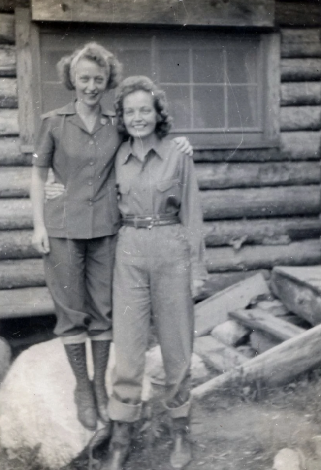 Two women in vintage military-style shirts and pants stand outdoors, smiling with arms around each other. They are in front of a rustic log building with a window. Rocks and wooden steps are visible at their feet.