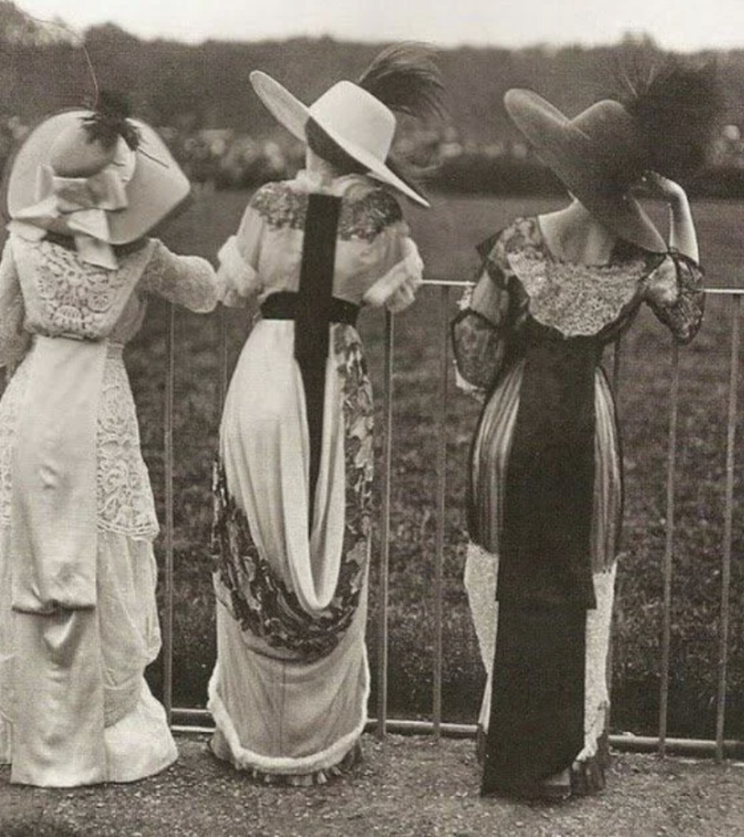 Three women dressed in Edwardian fashion, wearing large hats with plumes and intricate lace dresses, stand with their backs to the camera, observing an event behind a metal fence.