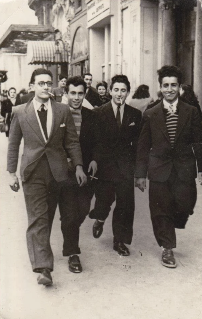 A vintage photo shows four young men in suits and ties walking confidently down a bustling city street. They are smiling, and the street is lined with people and buildings in the background. The photo appears to be from the mid-20th century.