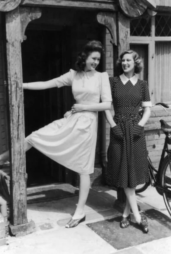 Two women in vintage dresses stand outside a brick building. One woman playfully lifts her leg and rests her foot on a wooden post, while the other smiles with her hands in her dress pockets. A bicycle leans against the wall nearby.