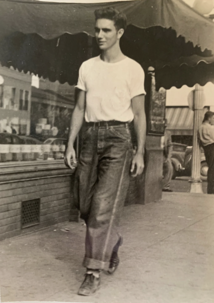 A young man in a white t-shirt and cuffed jeans walks casually on a city sidewalk. He is passing by a storefront with a large window display. The scene has a vintage, black-and-white aesthetic, suggesting a mid-20th century setting.