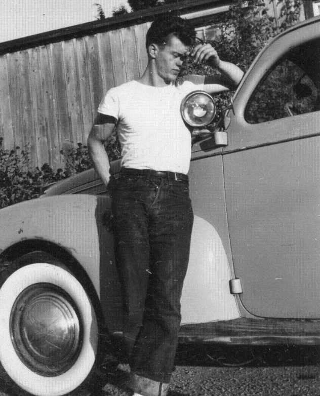 A man in a white T-shirt and jeans leans against a vintage car, resting his arm on the headlight. He appears contemplative, with his other hand touching his forehead. The background includes a wooden fence and foliage. The photo is in black and white.