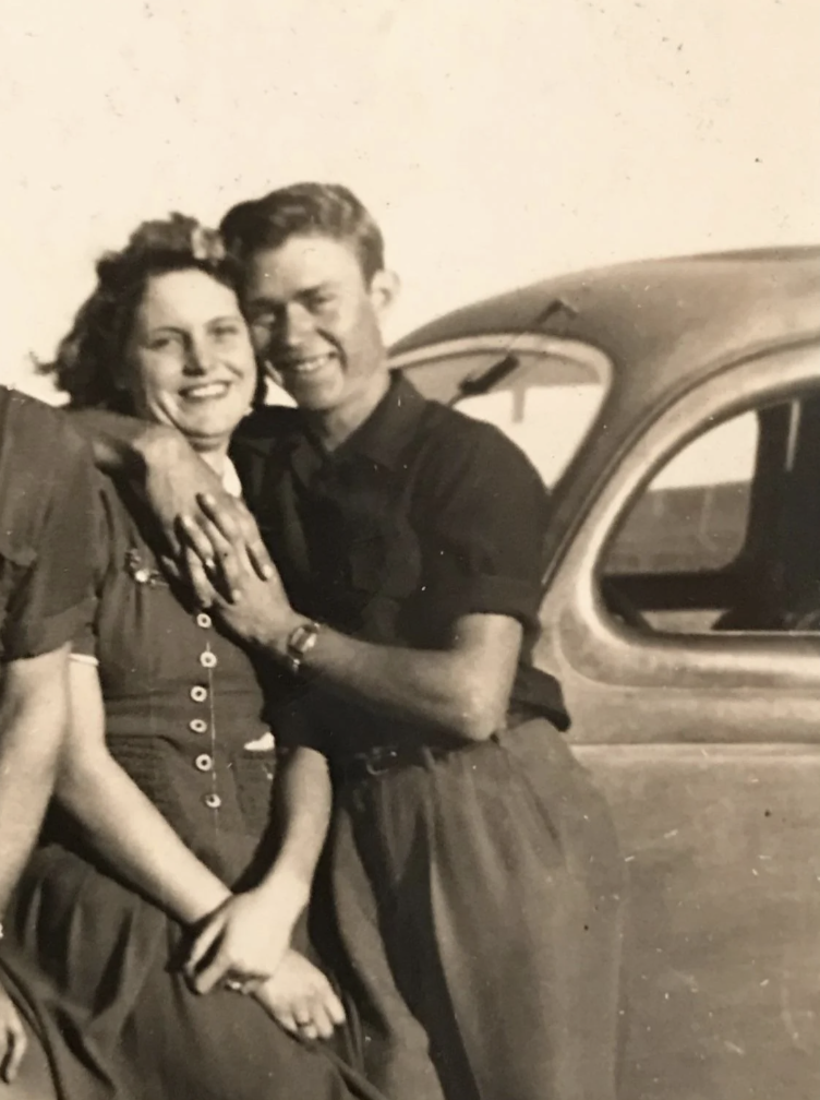 A smiling couple from the mid-20th century stands together in front of a vintage car. The man has his arm around the woman, who is wearing a button-down dress. Both are looking at the camera, radiating a sense of happiness and affection.