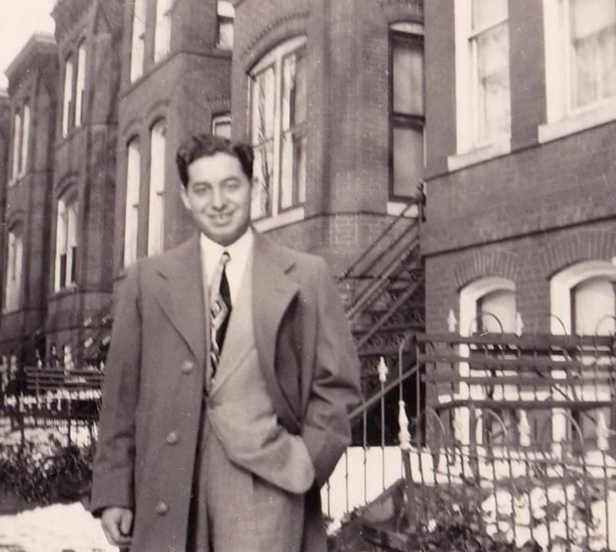 A man in a suit and tie stands smiling in front of a row of brick houses. He wears a long coat over his suit, and one hand is in his pocket. Snow is on the ground, and a decorative iron fence is in the foreground. The photo is in black and white.