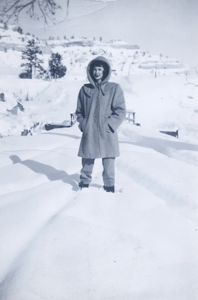 A person in a hooded coat stands in deep snow on a sunny day. The scene is a snowy landscape with trees and hills in the background. The person is smiling with hands in pockets, casting a shadow on the snow.