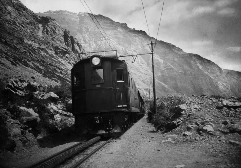 A black and white photo of a train traveling through a rocky mountainous landscape. The train is moving towards the camera on a narrow track, with overhead wires above. Steep cliffs rise dramatically in the background under a cloudy sky.