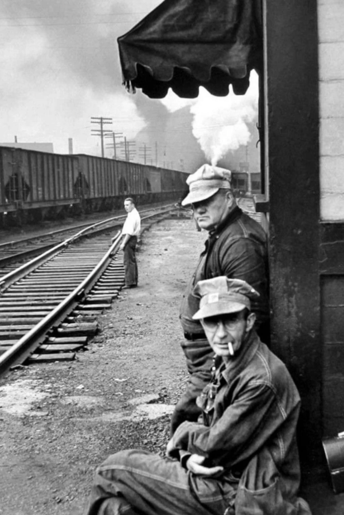 Three men are near train tracks, with two sitting close to a building and one standing by the rails. Smoke billows from a distant train. The setting appears industrial, with overcast skies and electrical poles visible in the background.