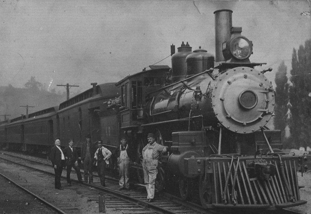 Black and white photo of a vintage steam locomotive on railroad tracks. Five men stand beside the train, wearing work attire and hats. The train has a prominent round front light and billowing smoke. Trees and utility poles are visible in the background.