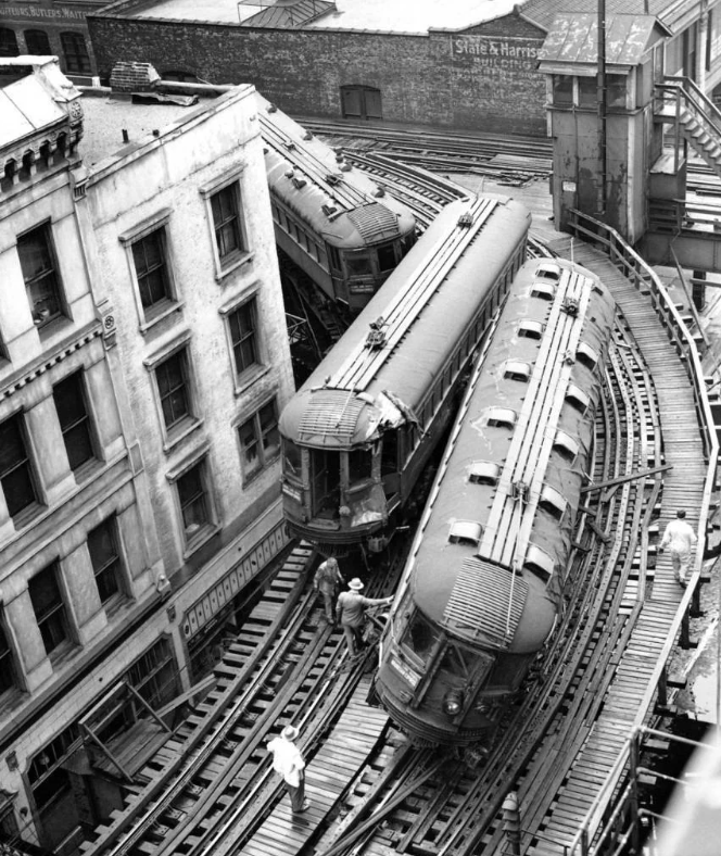 Black and white image of a vintage elevated train track with two trains rounding a curve between aged buildings. Men in work clothes stand on the tracks, appearing to inspect or carry out maintenance work.