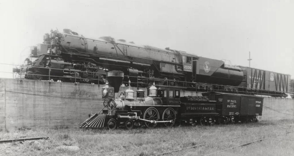 A black and white photo showing two trains: a small, antique steam locomotive in the foreground labeled "St. Paul & Pacific" and a large, modern train with the label "Union Pacific" on the elevated track behind it.