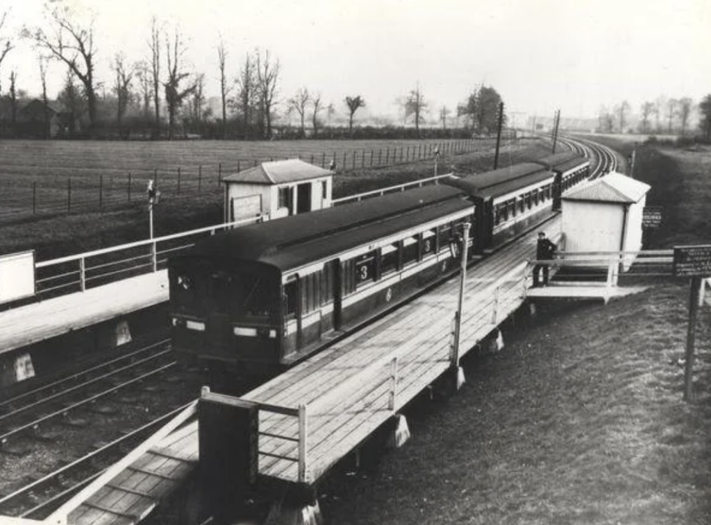 Black and white image of a train at a rural station, with wooden platforms and small buildings. The track curves into the distance, surrounded by fields and trees, under an overcast sky. A person stands near the train.