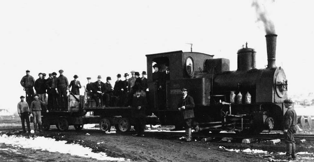 A vintage black and white photograph of a steam locomotive with several men standing on and around it. The train is on a dirt track, with some snow patches visible on the ground. Smoke rises from the train's smokestack. The men are dressed in early 20th-century attire.