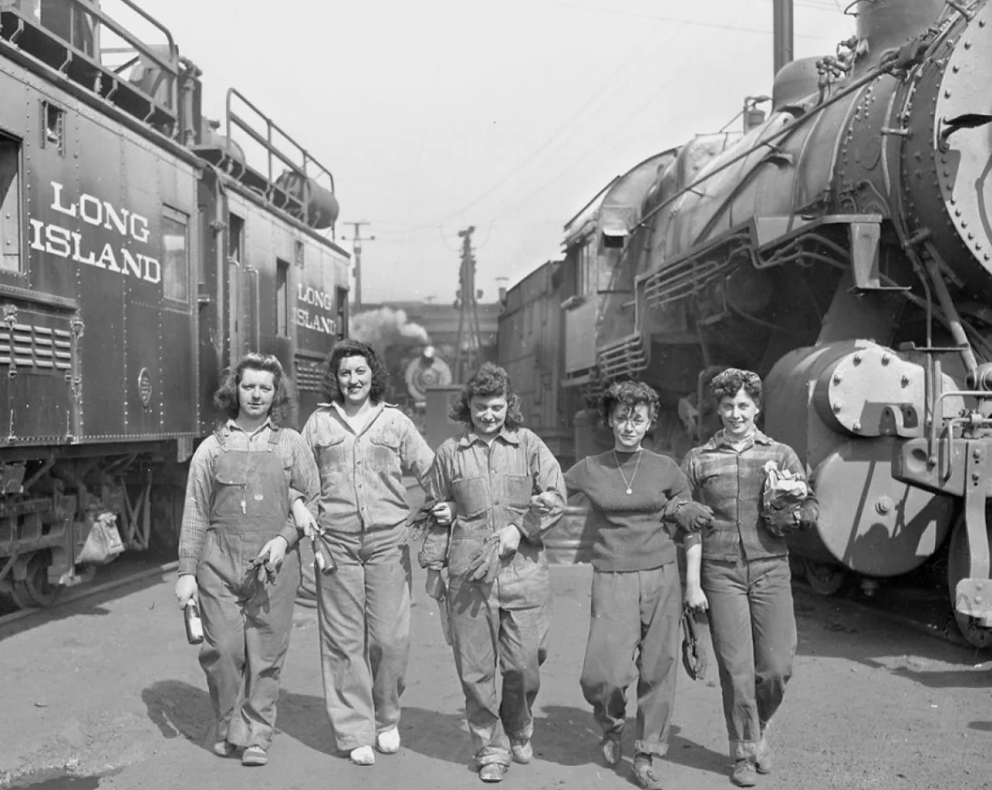 Five women walk arm in arm beside steam locomotives labeled "Long Island." They wear coveralls and work attire, suggesting involvement in railway operations. One woman holds a bottle, while another carries a helmet. The scene is black and white.