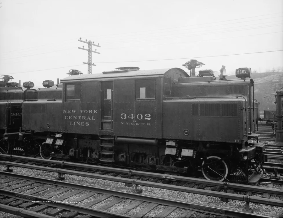 Black and white photo of a New York Central electric locomotive, numbered 3402, on railway tracks. The engine has a boxy design and visible pantographs. Power lines and other trains are in the background, with a cloudy sky overhead.