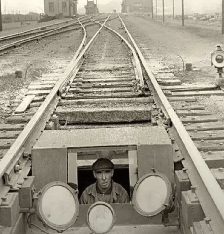 A man wearing a cap is seen inside a maintenance cart on railway tracks. The cart is positioned between two tracks that split in the background. The setting appears industrial, with buildings and other rail equipment visible.