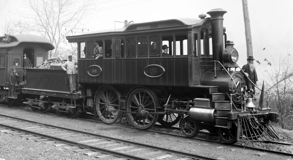 A black and white photo of an old steam locomotive with large wheels and a tall smokestack. People stand by the train, with some on the coal carriage. Sparse trees and an overcast sky are in the background.