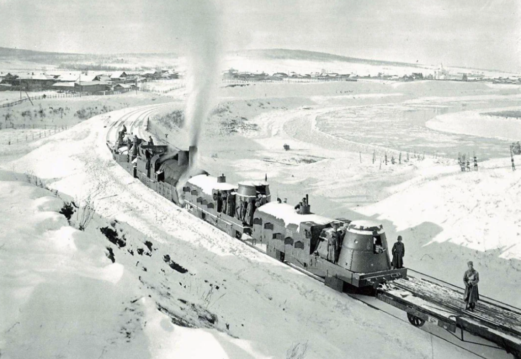 Steam locomotive plows through a snowy landscape with smoke billowing from its chimney. Two people stand on a flatbed train car. The scene is set against a backdrop of snow-covered hills and buildings in the distance.