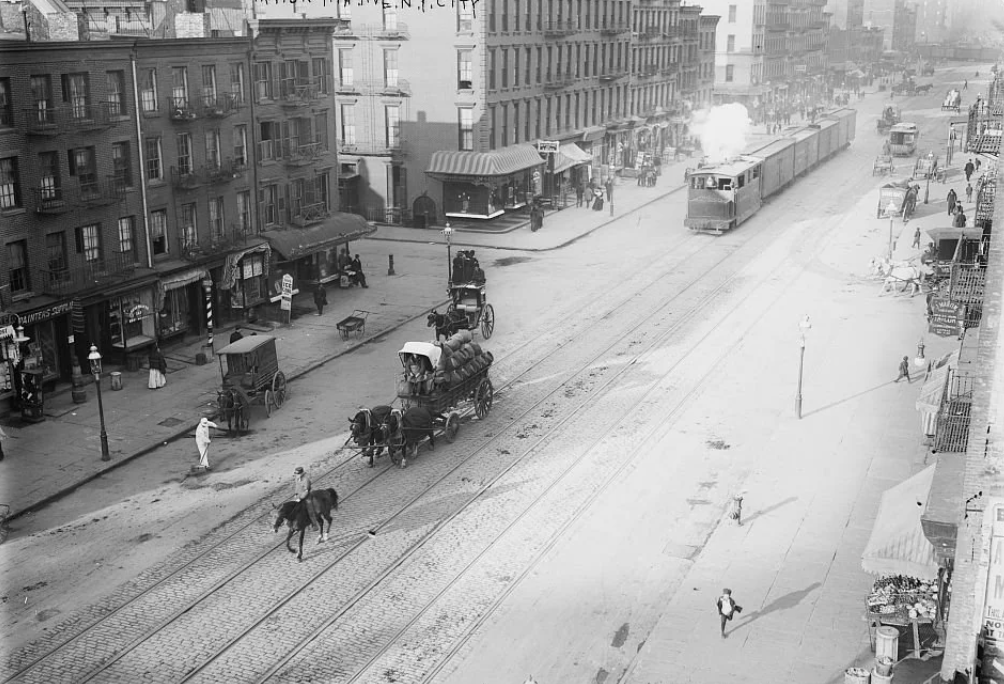 A historic black-and-white photo shows a busy early 20th-century city street. Horse-drawn carriages, a streetcar, and several pedestrians are visible. Buildings line both sides of the street, and shops are on the ground level.