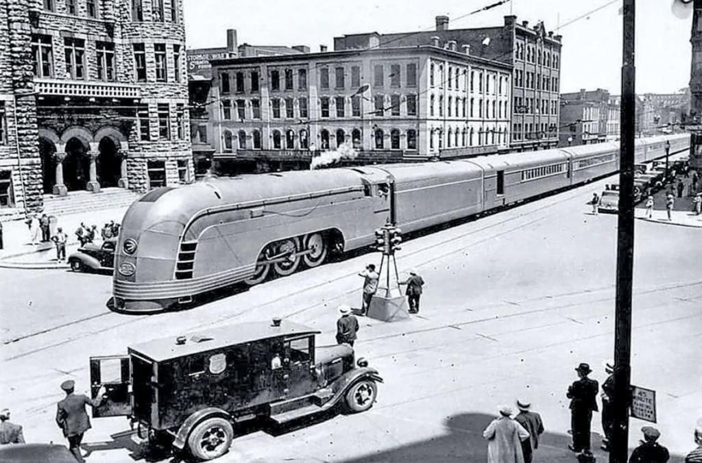 A historic black and white photo depicts a streamlined, Art Deco-style train passing through an urban area. The street has vintage cars and people dressed in early 20th-century attire.