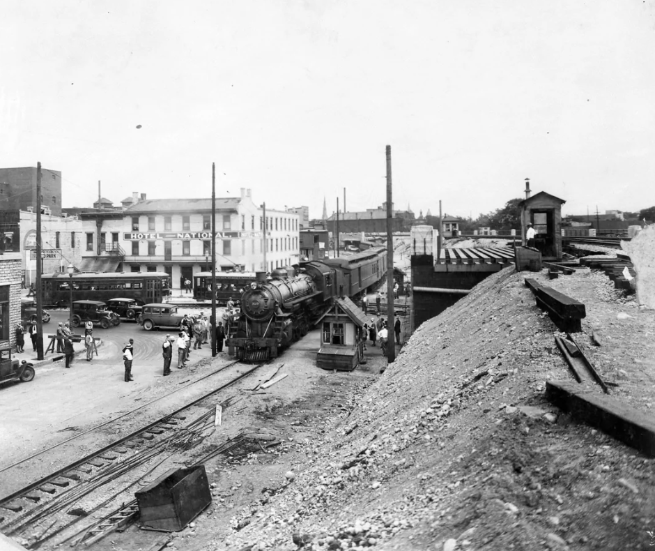 A vintage black and white photo of a steam locomotive at a railroad crossing in a town. People gather nearby, with cars and buildings in the background, including a sign for a hotel. The foreground shows a gravelly embankment and tracks.