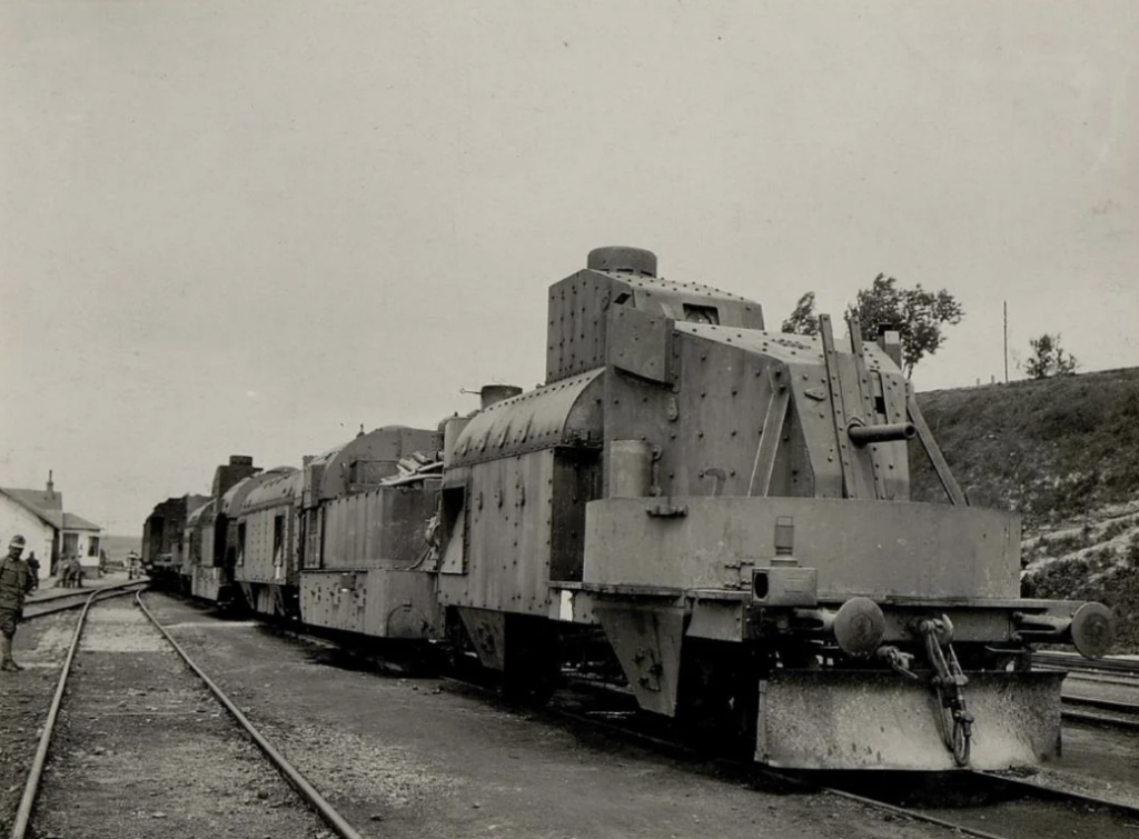 A vintage photo of an armored train with metal plating, standing on railway tracks. The train has a robust, fortified appearance. A figure stands near the track, and there are buildings and foliage in the background.