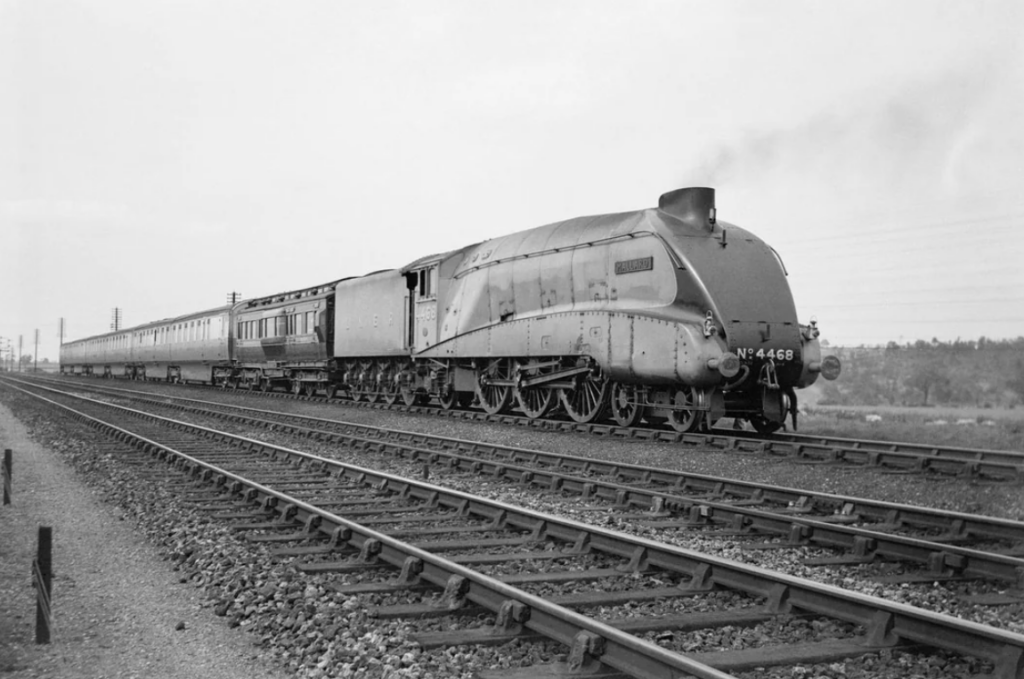 Black and white photo of a streamlined steam locomotive with a distinctive aerodynamic design, pulling a train of passenger carriages. The train is traveling on a set of parallel tracks in an open area.