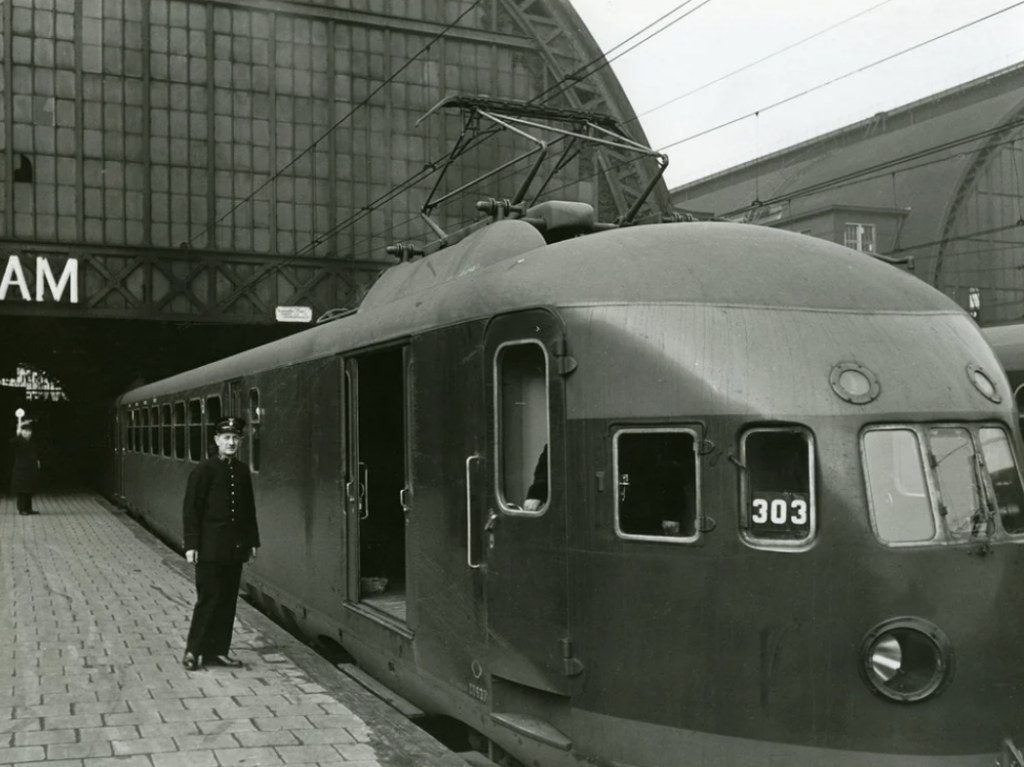 A black and white photo of a historic train with the number 303 at a station platform. A conductor in uniform stands beside the open train door. The background shows a large building with glass and steel architecture, partially displaying the word "AM.