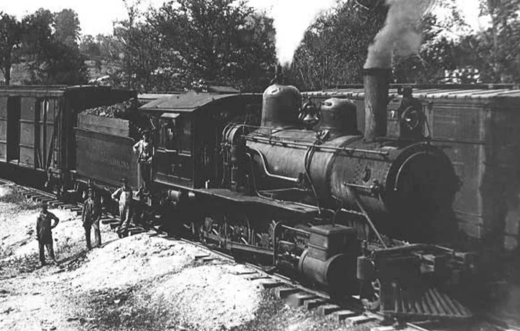 Black and white photo of a vintage steam locomotive on tracks, emitting smoke. Two men stand on the train, while three others stand beside it. Trees are in the background, suggesting a rural setting.