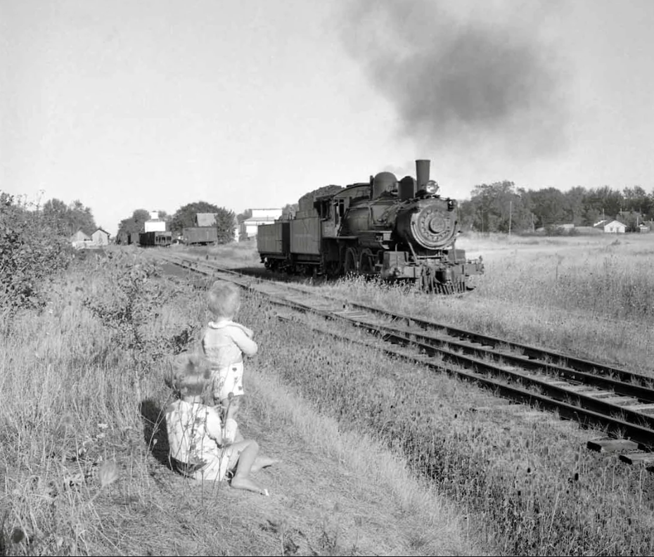 Black and white photo of two children sitting beside a railroad track, watching a steam locomotive approaching from a distance across a grassy field with scattered trees and buildings in the background.