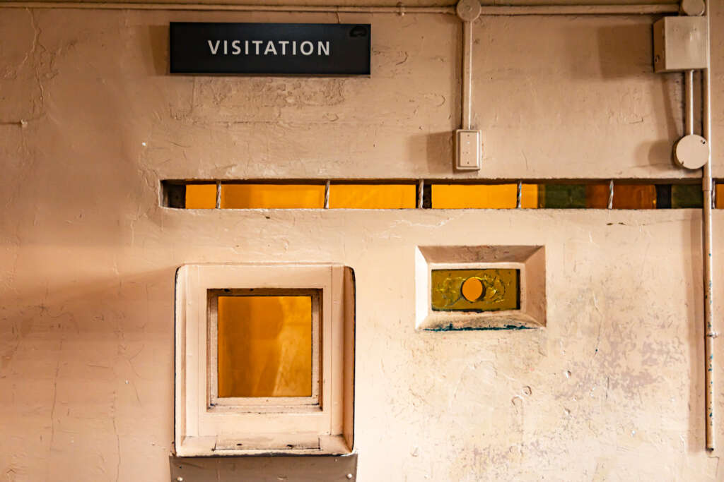 A textured wall with a small square window, surrounded by stained glass accents. Above, a black sign with white text reads "VISITATION." The setting appears aged and institutional.