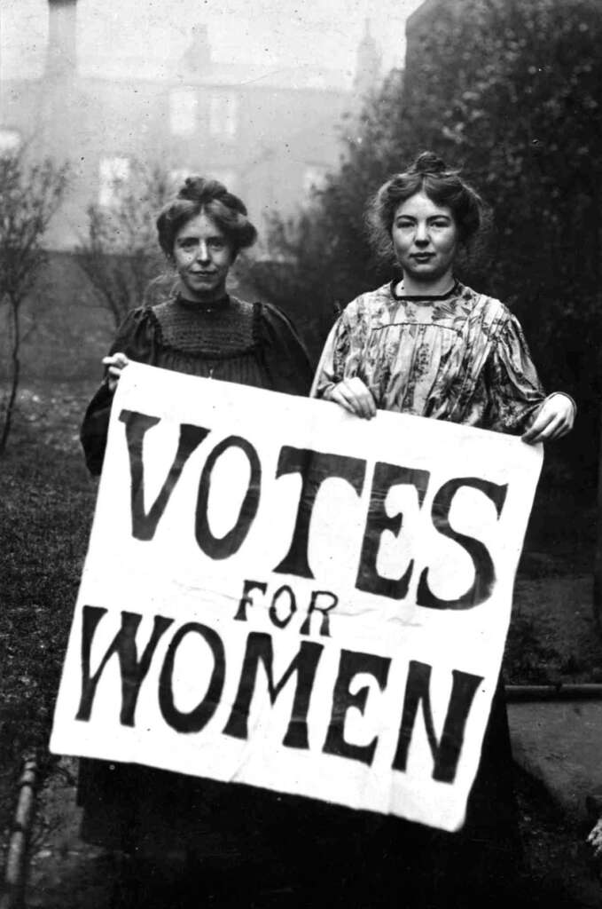 Two women stand outdoors holding a large sign that reads "Votes for Women." Both wear early 20th-century clothing. The background is slightly blurred with trees and a building visible.