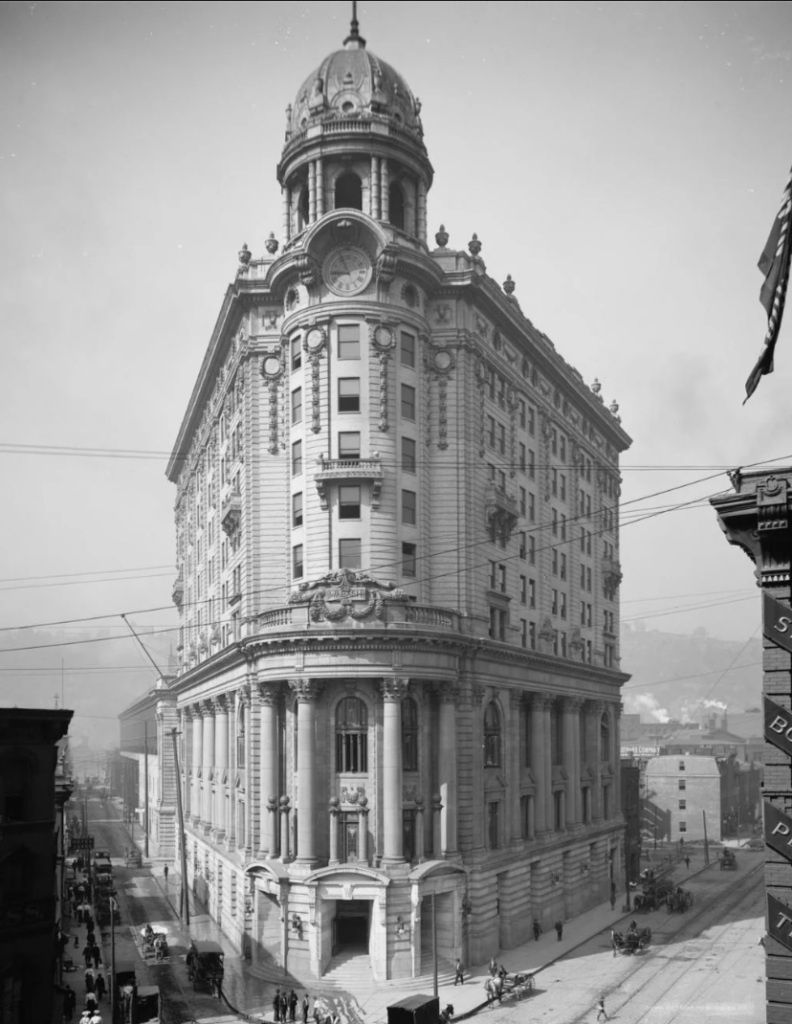 Black and white image of a historic multi-story building with ornate architecture, featuring a dome and clock at the top. The street below is bustling with people and horse-drawn carriages. Other buildings and a hilly background are visible.