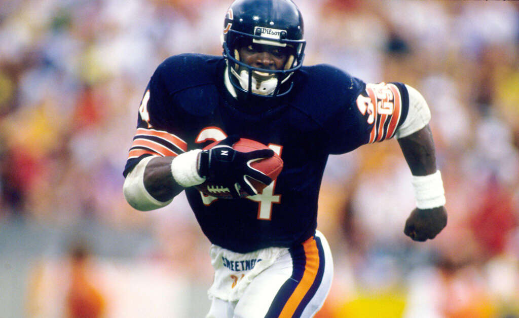 A football player in a navy blue and orange uniform runs with the ball on a field. The jersey has the number 34, and he wears a helmet with a face mask. The background shows a blurred crowd.