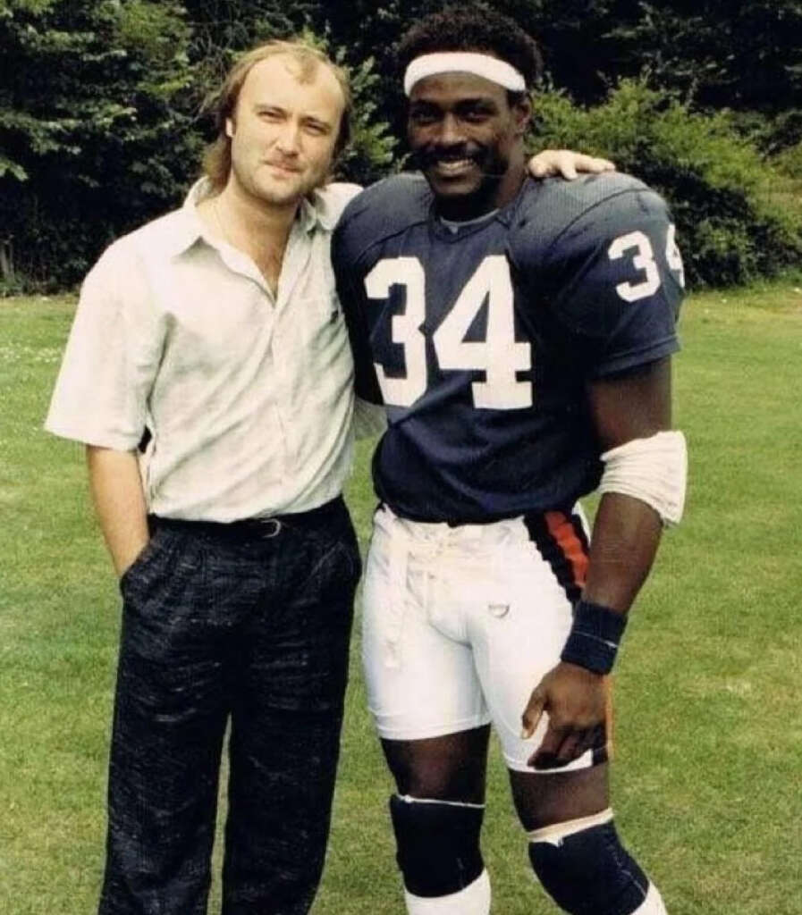 Two people are standing together outdoors. One is wearing casual clothing, and the other is in a navy and orange football uniform with number 34. Both are smiling, with trees and grass in the background.