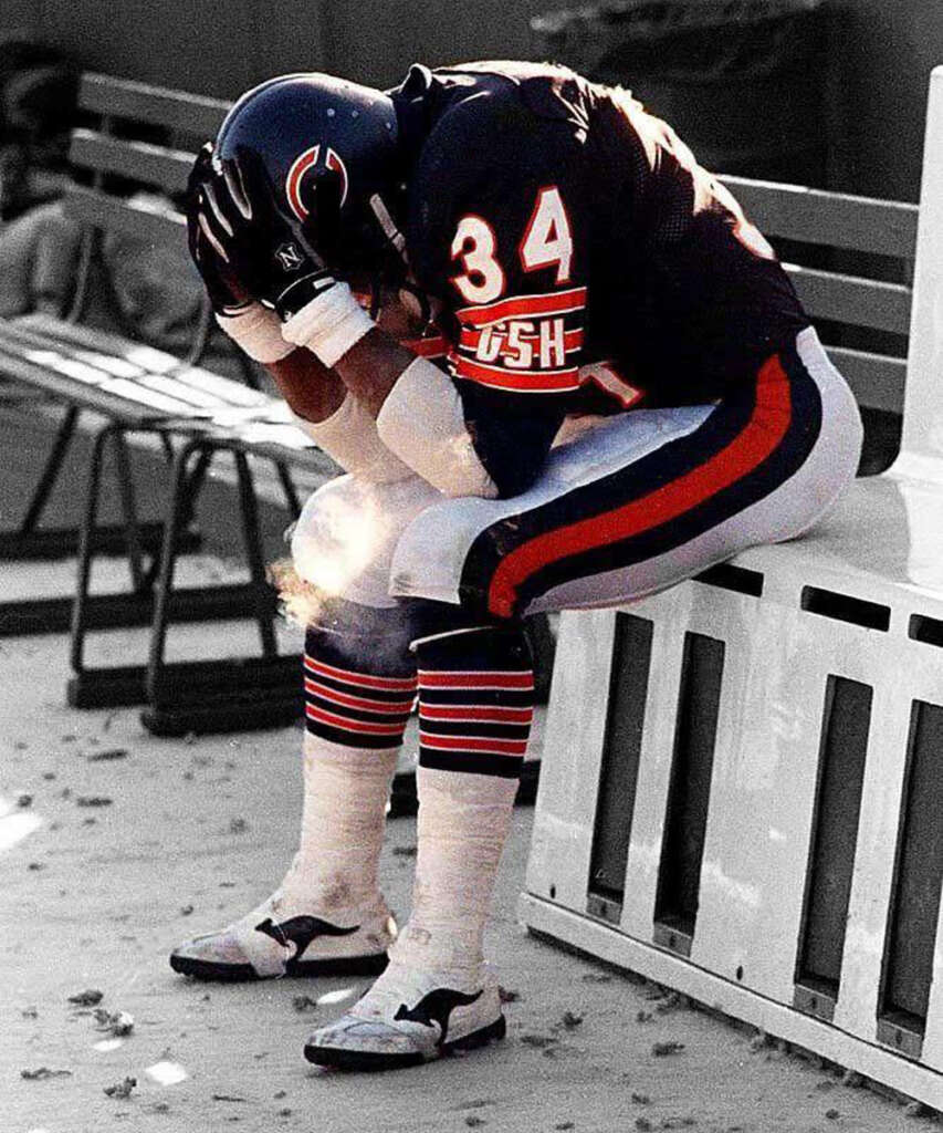 American football player in a dark uniform, number 34, sits on a bench with head in hands, appearing dejected. The background is a sports field with scattered leaves on the ground.