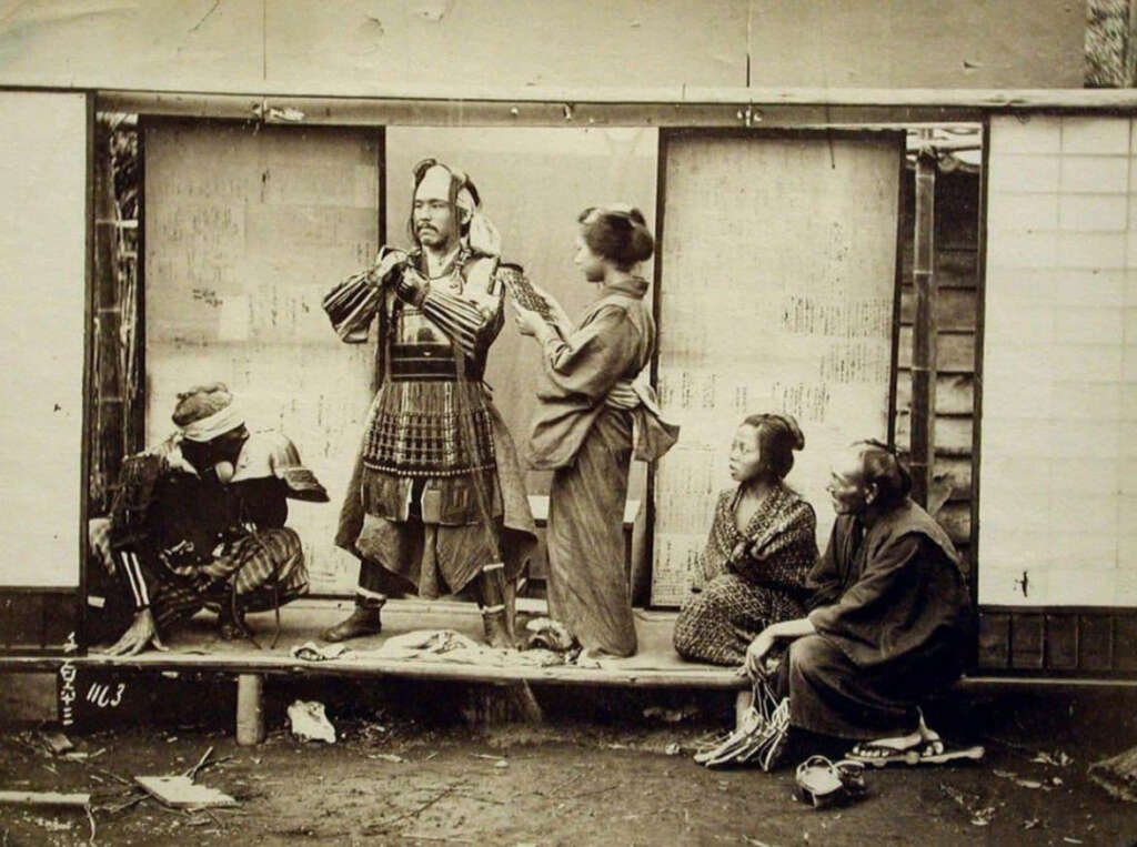 A historical photograph shows a man in samurai armor being assisted by three women in traditional Japanese attire. Another man, also in traditional clothing, sits nearby. They are on a wooden platform with a backdrop of sliding paper doors.