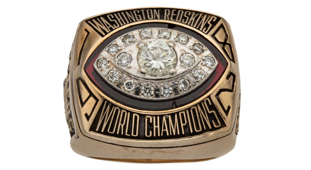 A gold championship ring with "Washington Redskins" and "World Champions" engraved. The center features a football shape adorned with diamonds, including a large central diamond, surrounded by smaller ones.