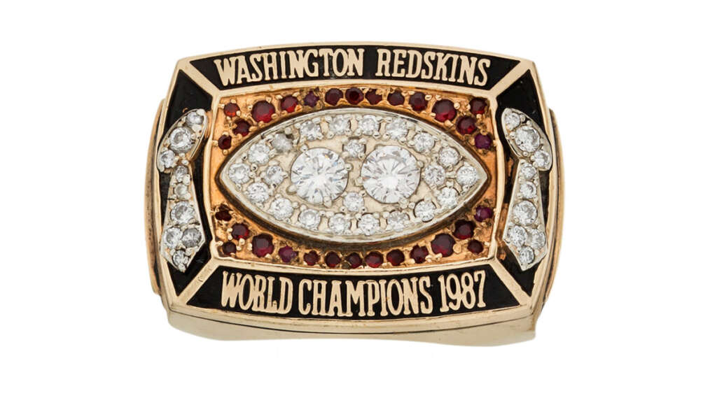 A gold championship ring with the text "Washington Redskins" on the top and "World Champions 1987" on the bottom. The center features three large diamonds surrounded by smaller red and white stones in a football shape.