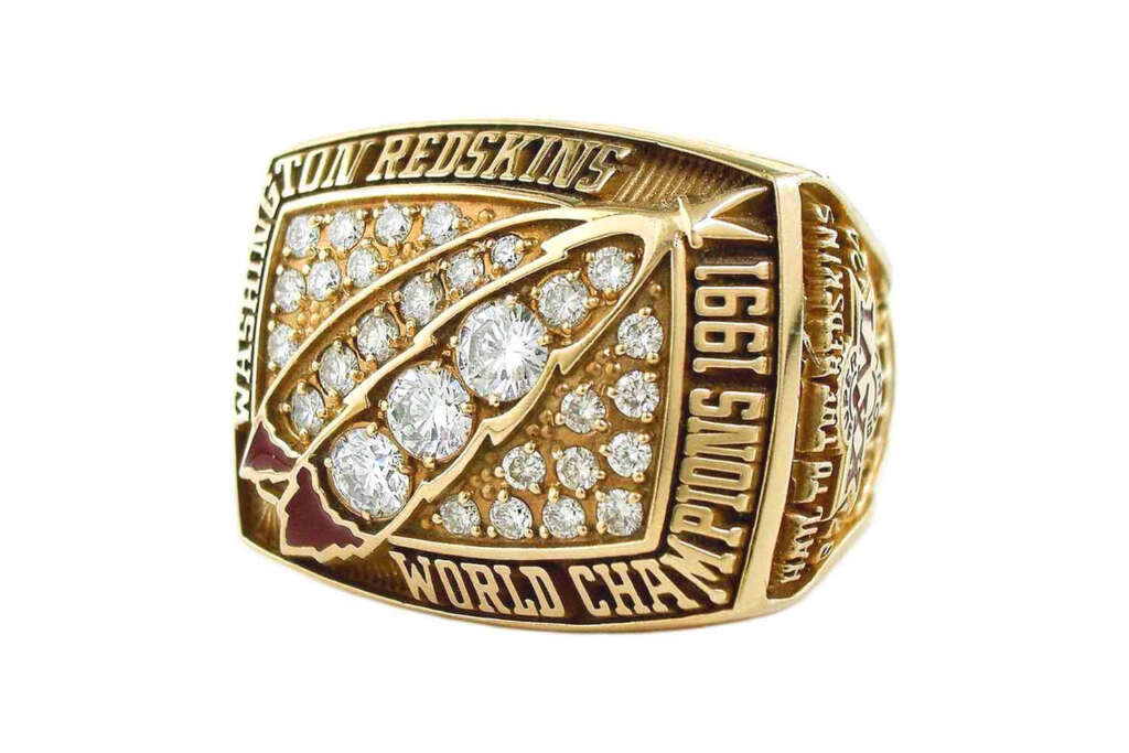 Gold championship ring with "Washington Redskins" and "World Champs" engraved. Features a football design and "Super Bowl XXVI 1991" text adorned with numerous diamonds.