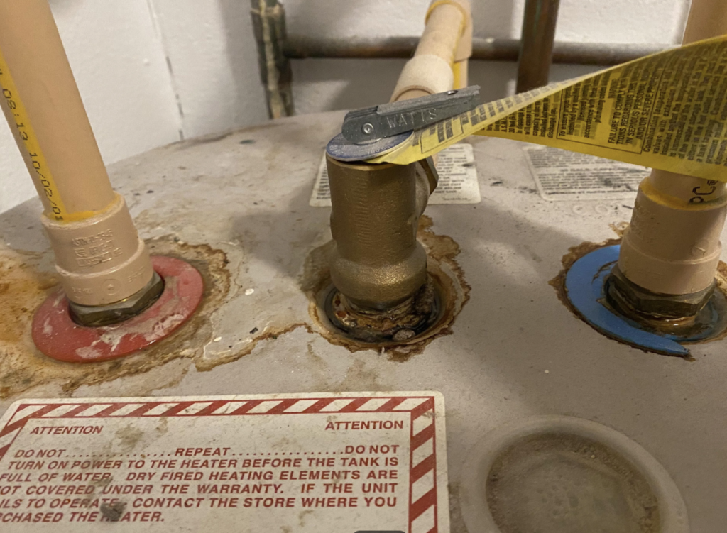 Close-up of a water heater with corroded pipes and fittings. A wrench labeled "WATTS" rests on one pipe. A warning label is visible with several red and black texts. Surrounding pipes show rust and signs of wear.