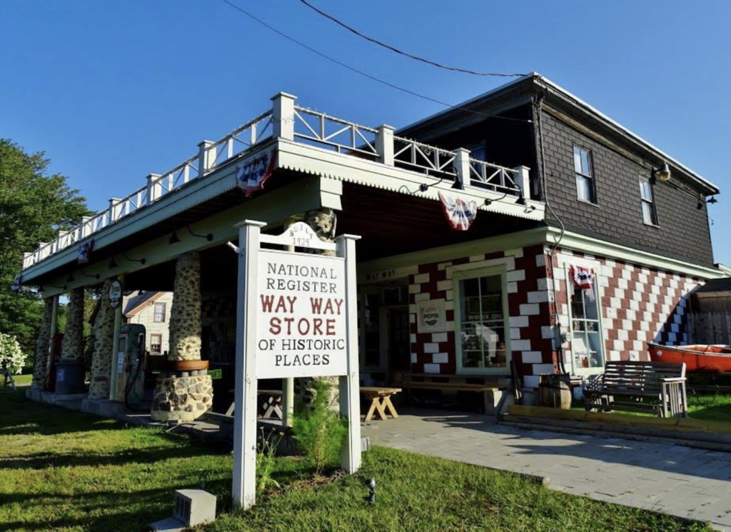 A historic store with a sign reading "National Register Way Way Store of Historic Places." The building has a red and white brick pattern, a porch with seating, and decorative flags. A grassy lawn and a tree are in the foreground.