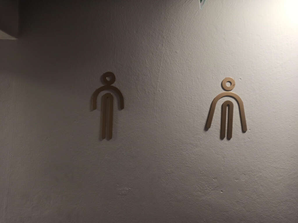 Wall with two abstract restroom symbols. The left symbol suggests a male figure, and the right symbol implies a female figure. Both are made from simple lines and circles on a textured gray wall.