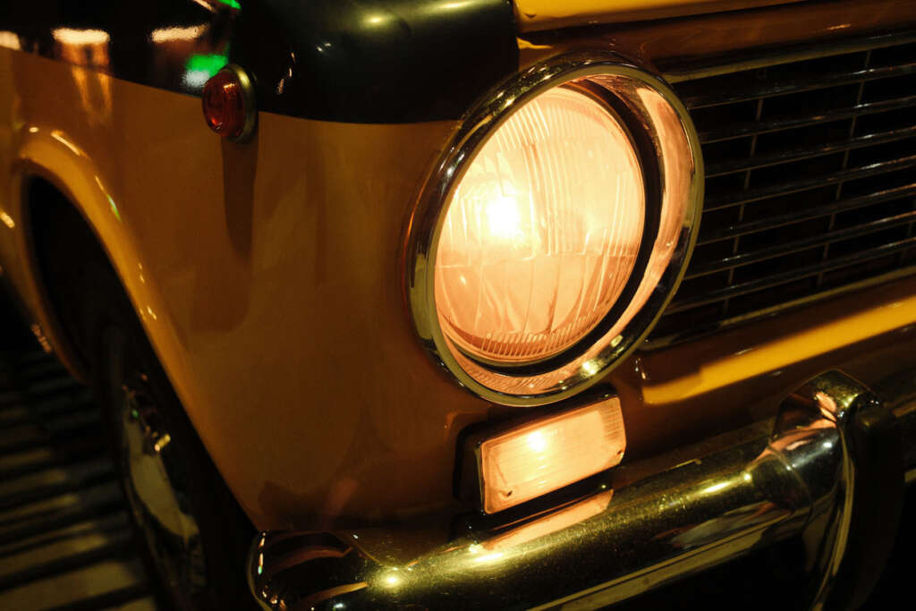 Close-up of a vintage car's front, featuring a round headlight illuminated in a warm glow. The car is yellow with dark trim, and part of the grille and bumper are visible. The scene appears to be lit by ambient light.