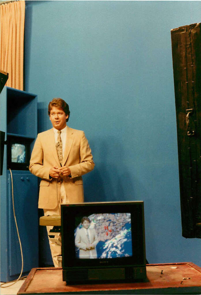 A man in a beige suit stands in a television studio, presenting the weather. A monitor in front shows the same image with a highlighted weather map. The background is a solid blue, and studio equipment is visible around him.